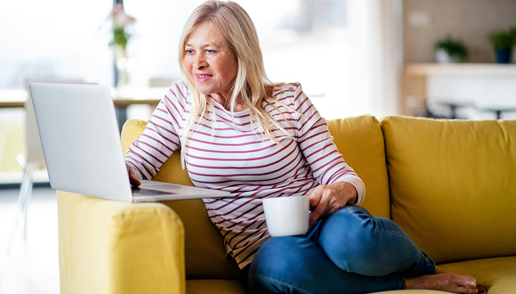 middle aged woman sitting on coach with laptop and coffee cup